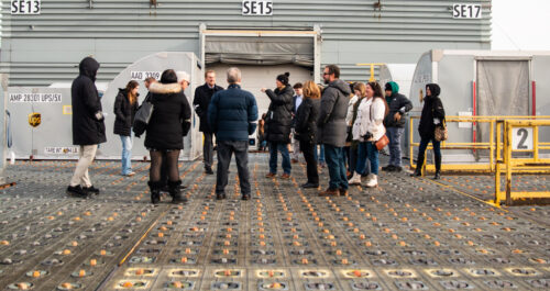 A group of people wearing winter jackets stand on a grid-like surface in front of a large industrial structure with signs reading "SE15" and "SE17." The ground features circular patterns, and a UPS container hints at the upcoming UPS Event.