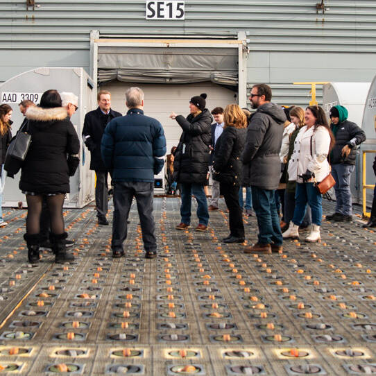 A group of people wearing winter jackets stand on a grid-like surface in front of a large industrial structure with signs reading "SE15" and "SE17." The ground features circular patterns, and a UPS container hints at the upcoming UPS Event.