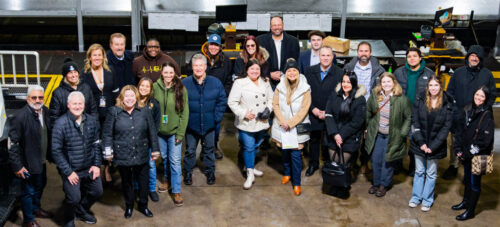 A group of people, dressed warmly in coats and jackets, stand together at the 2024 UPS event in an industrial setting. They're smiling and posing for the photo, surrounded by equipment and machinery in the background.
