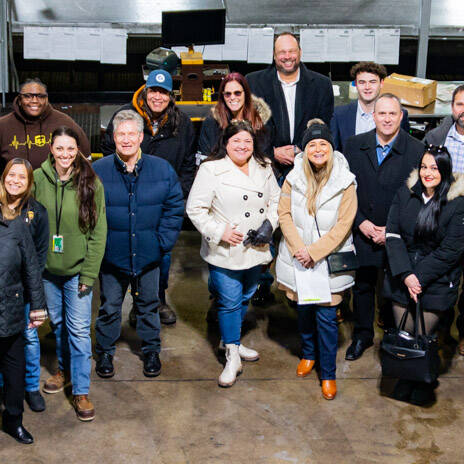 A group of people, dressed warmly in coats and jackets, stand together at the 2024 UPS event in an industrial setting. They're smiling and posing for the photo, surrounded by equipment and machinery in the background.