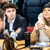 At the UPS event, a group of people sit around a table. A young man in a suit and a woman in a winter coat and beanie are at the forefront, engaged in conversation. A handbag and water bottles are on the table, adding to the casual yet professional atmosphere.