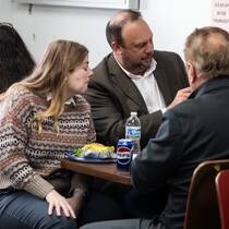 A group of people sit around a table at the UPS Conference, engaged in lively conversation. One person is holding a smartphone, while others enjoy drinks and snacks. The setting appears casual, emphasizing interaction and collaboration.