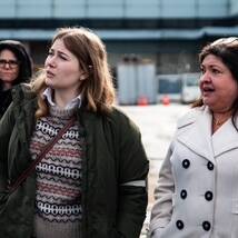 Three people stand outdoors in front of a building, possibly during the 2024 UPS event. A person in a dark jacket smiles on the left, while the person in the center wears a green jacket and looks ahead. The person on the right, in a white coat, also looks forward with a slight smile.