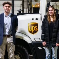 In a garage, two individuals stand before a white UPS truck, its logo clearly displayed. The person on the left sports a suit jacket, while the one on the right dons a black coat. This scene is set against the backdrop of an upcoming 2024 UPS event.