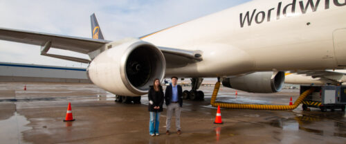 A man and woman stand on a wet airport tarmac beside the engine of a large UPS cargo plane, with "Worldwide" visible on the side. Orange cones are strategically placed around the area during this 2024 UPS event under an overcast sky.