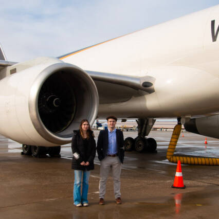 A man and woman stand on a wet airport tarmac beside the engine of a large UPS cargo plane, with "Worldwide" visible on the side. Orange cones are strategically placed around the area during this 2024 UPS event under an overcast sky.