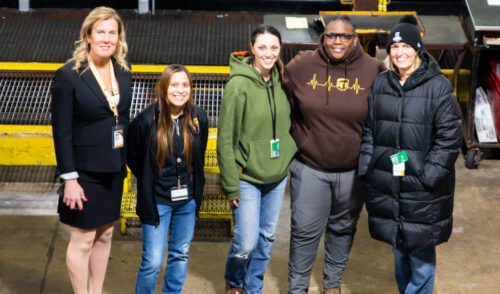 Five people are standing together in a warehouse setting at the UPS Event 2024. They are wearing a mix of casual and professional attire, including jackets and a sweatshirt with a heart monitor graphic. They're smiling and have ID badges hanging from lanyards.