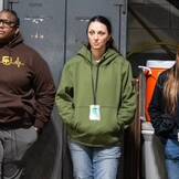 Three people stand in a room with industrial equipment at the UPS Event 2024. One wears a brown hoodie, another a green jacket, and the third a black jacket. They all have lanyards and appear to be listening attentively.