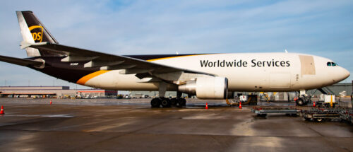 A UPS Worldwide Services cargo airplane is parked on the airport tarmac beneath a clear 2024 sky. The aircraft, adorned in white with brown and yellow stripes, stands ready with cargo equipment nearby, set to support another essential global event.
