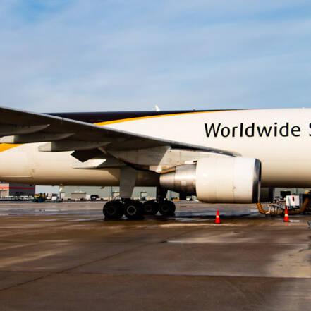 A UPS Worldwide Services cargo airplane is parked on the airport tarmac beneath a clear 2024 sky. The aircraft, adorned in white with brown and yellow stripes, stands ready with cargo equipment nearby, set to support another essential global event.