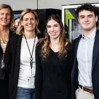 Four people, three women and one man, stand smiling in front of vending machines at the 2024 UPS event. Two women wear black blazers, another wears a black top, and the man is dressed in a navy blazer and khaki pants.