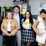 A group of eight people, dressed in winter and business casual attire, smiles at the camera indoors during a UPS 2024 event. One wears a checked skirt and another proudly sports a shirt with a green logo. The backdrop includes a wall and a vending machine, adding to the lively setting.