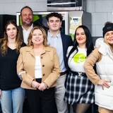 A group of nine people stands together indoors at the UPS Event 2024, all smiling. Some are dressed casually, while others wear business attire. The background features vending machines and posters on the wall, capturing the vibrant atmosphere of this significant gathering.