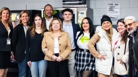 A group of nine people stands together indoors at the UPS Event 2024, all smiling. Some are dressed casually, while others wear business attire. The background features vending machines and posters on the wall, capturing the vibrant atmosphere of this significant gathering.