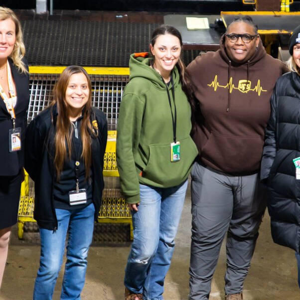 Five people are standing together in a warehouse setting at the UPS Event 2024. They are wearing a mix of casual and professional attire, including jackets and a sweatshirt with a heart monitor graphic. They're smiling and have ID badges hanging from lanyards.